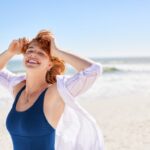Beautiful woman with red hair and freckles having fun in blue swimsuit on tropical beach, with copy space. Curvy young woman smiling and looking at camera at sea. Joyful girl in swimwear enjoying.