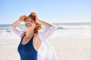 Beautiful woman with red hair and freckles having fun in blue swimsuit on tropical beach, with copy space. Curvy young woman smiling and looking at camera at sea. Joyful girl in swimwear enjoying.